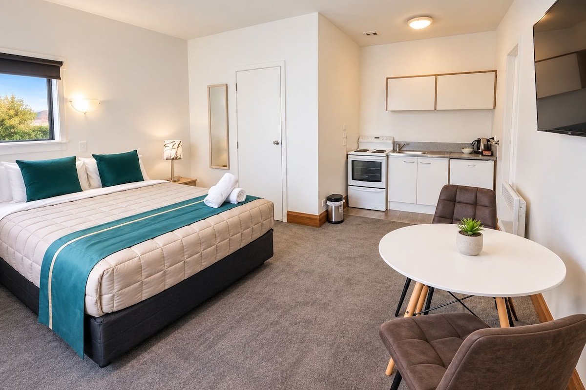A compact hotel room with a bed, a small kitchenette, a dining table, and a chair.