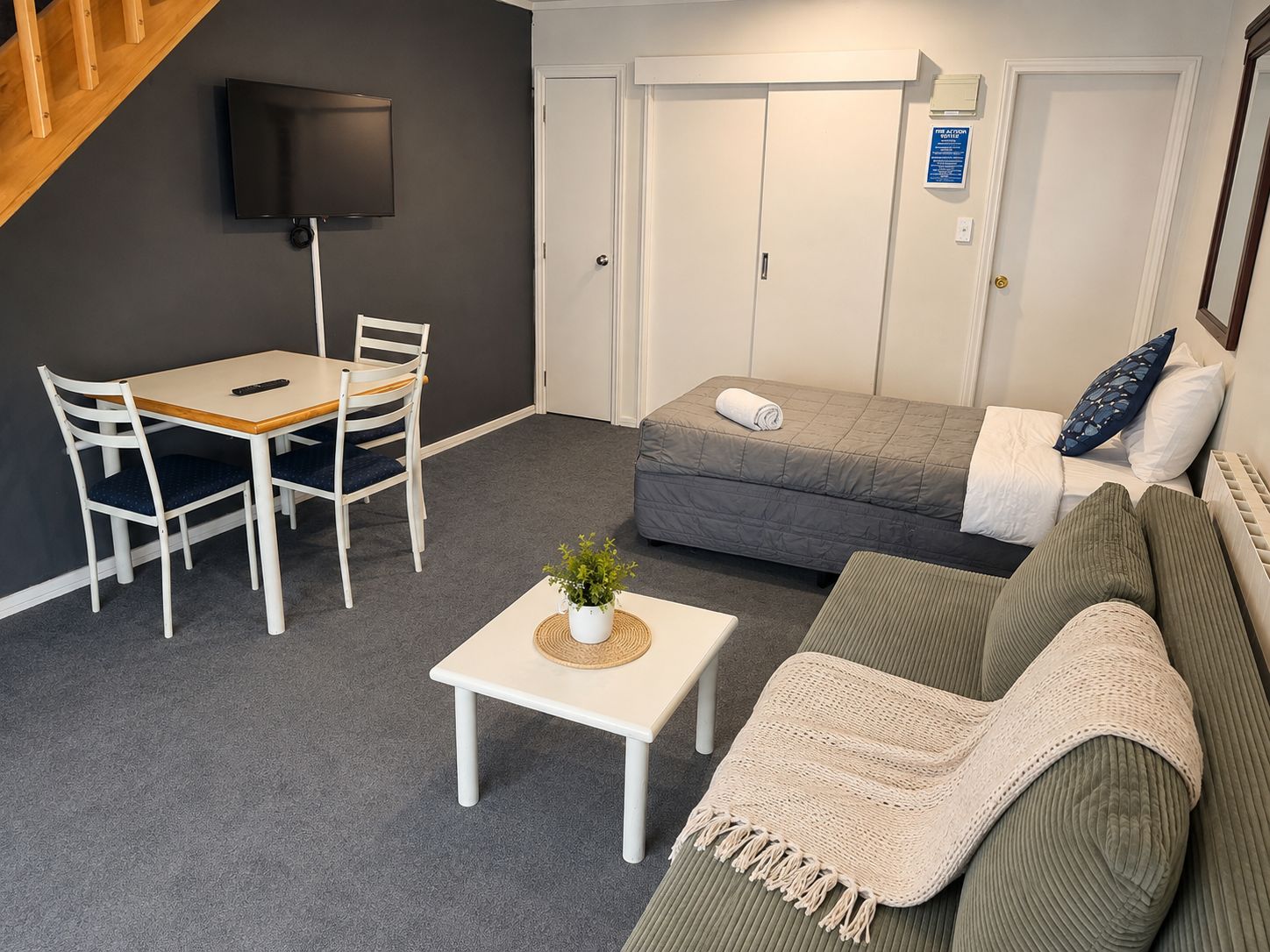 A compact living space with a sofa, a small table with a plant, a TV mounted on the wall, and a bed with pillows.