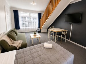 A modern living room with a sofa, a small table with a plant, a bed, a dining area with chairs, a staircase, and a TV mounted on the wall.