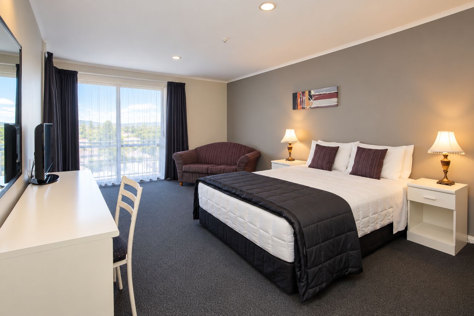 A hotel room with a bed, a desk, a chair, a sofa, two bedside tables with lamps, and a window with curtains.