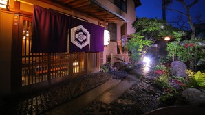 Un edificio japonés tradicional con fachada de madera, un sendero de piedra y un jardín con rocas y plantas.
