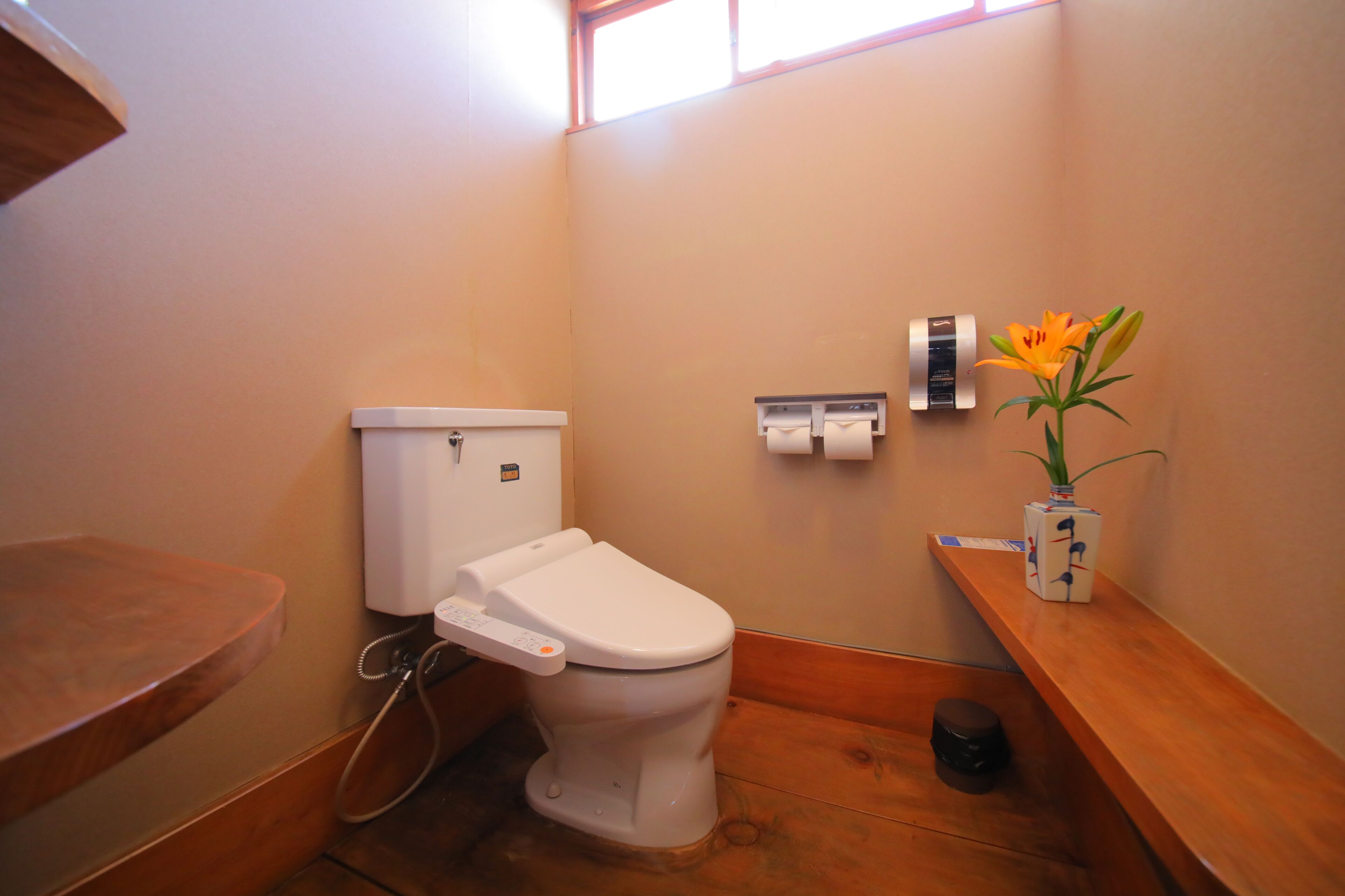Ein Badezimmer mit Toilette, einem Holzbodenregal, einem Toilettenpapierhalter und einer Vase mit Blumen.