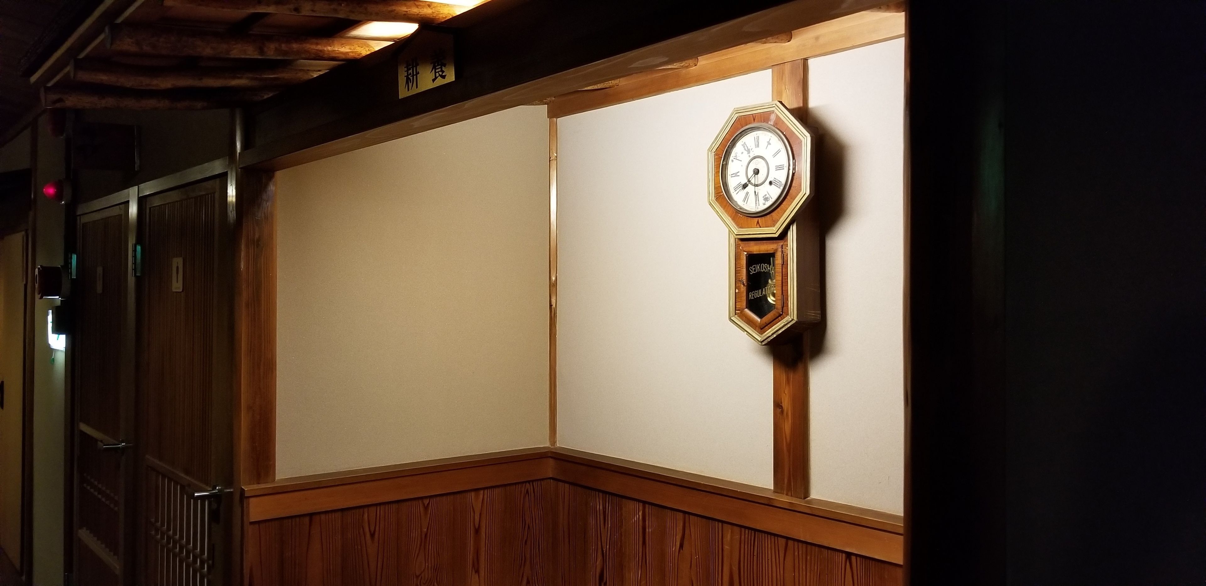 Esquina de una habitación con techo de madera, un reloj en la pared y una puerta con un cartel encima.