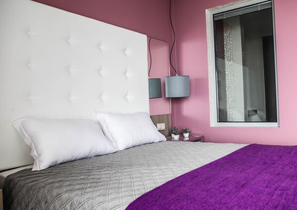 Un dormitorio con un cabecero capitonado, una cama con ropa de cama blanca y gris, una manta morada, una mesita de noche con dos plantas en macetas y una ventana con vistas a una pared de azulejos.