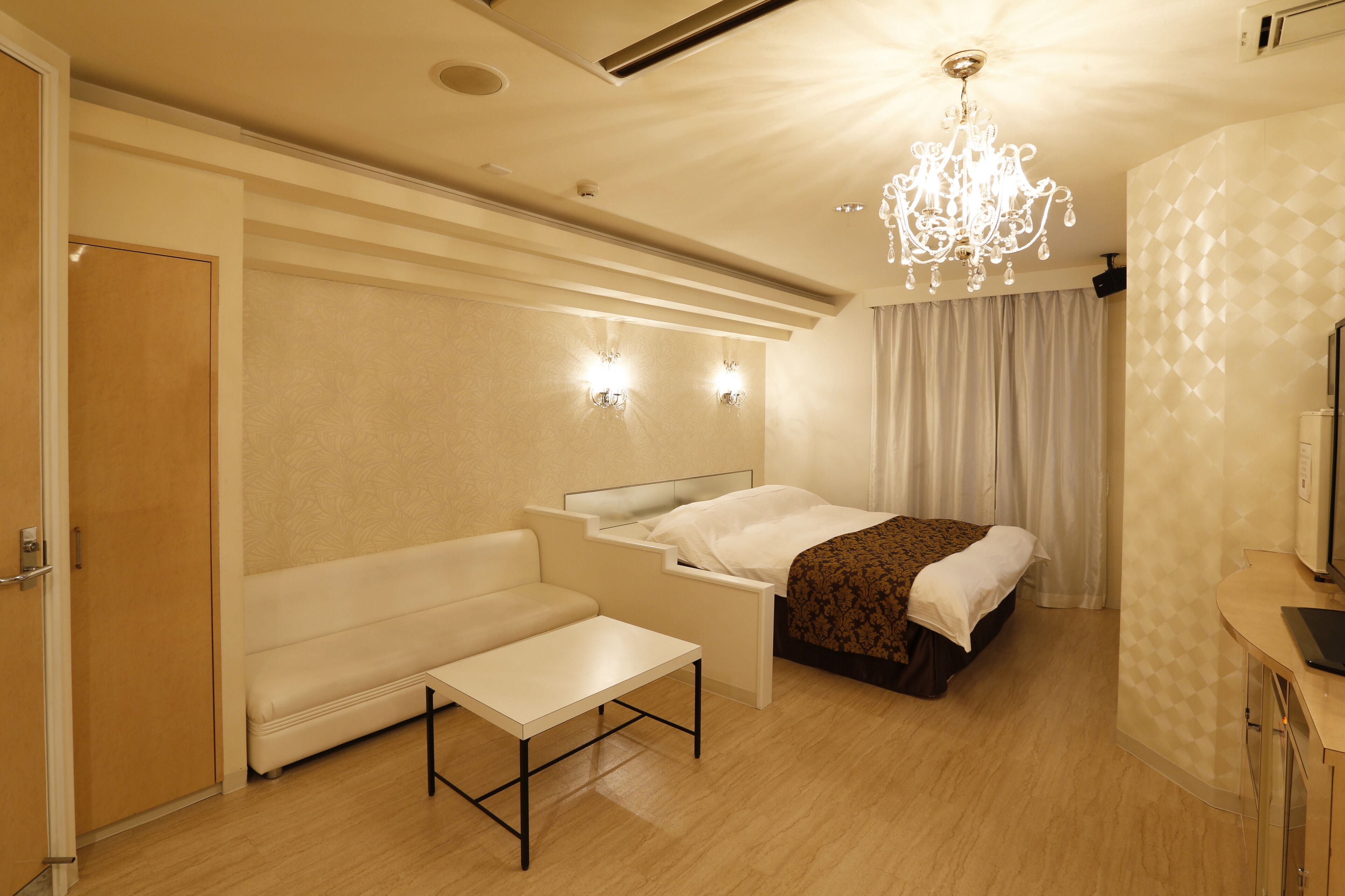 A hotel room with a bed, a bench, a small table, a mirror, and a chandelier.