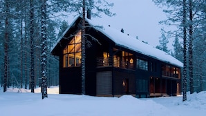 Une cabane dotée de grandes fenêtres et d’un toit en pente, entourée d’arbres couverts de neige.