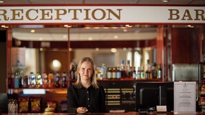 A reception area with a bar in the background, a counter with a cash register, and various bottles and glasses on shelves. - Hotel Savoy (Mariehamn)