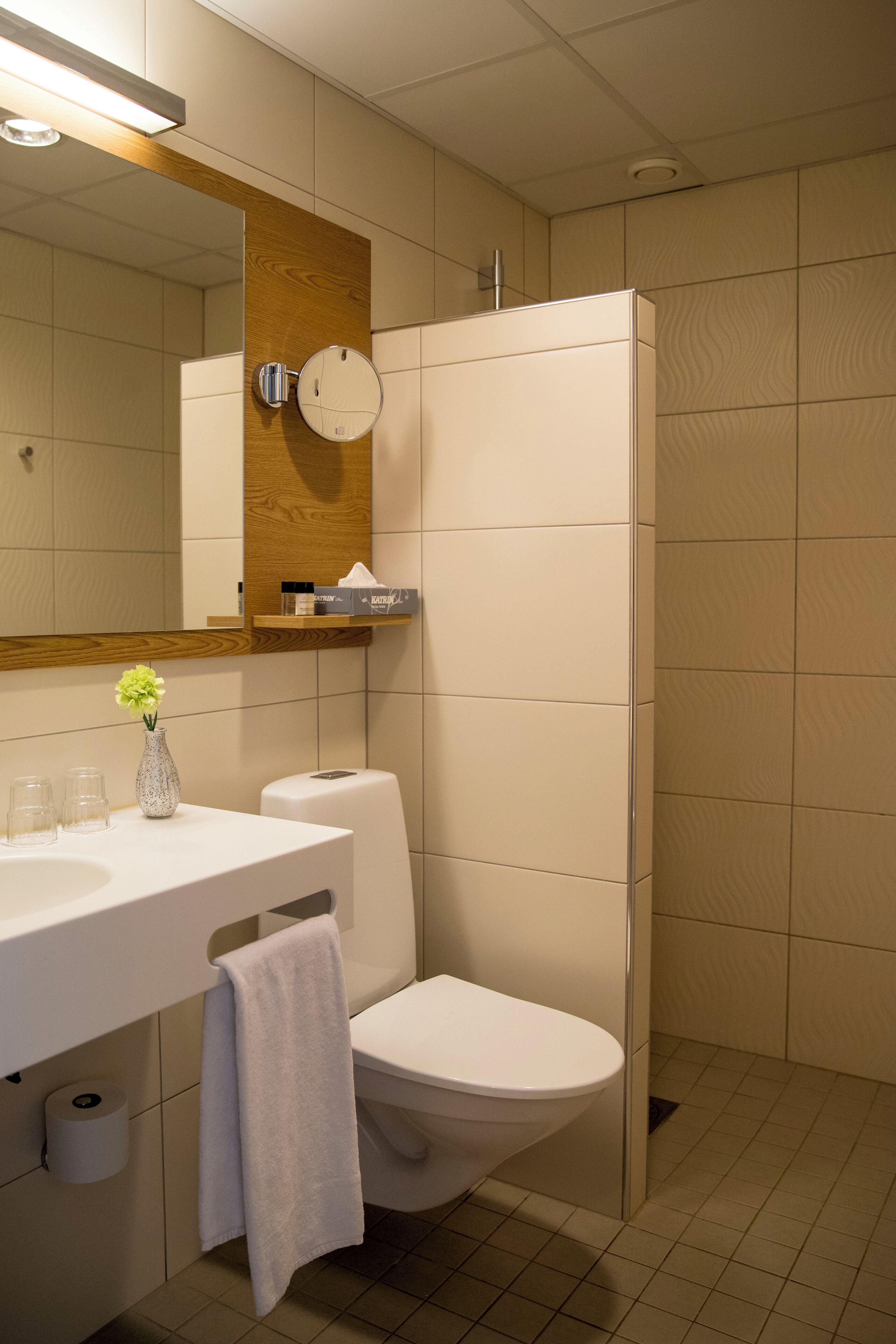 A modern bathroom with a white sink, mirror, and toilet. There is a towel hanging on the wall, a small vase with flowers, and a shelf with toiletries.