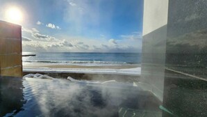 Uitzicht op een strand door een glazen deur met houten frame, leidend naar een kamer met een groot raam met uitzicht op de oceaan.