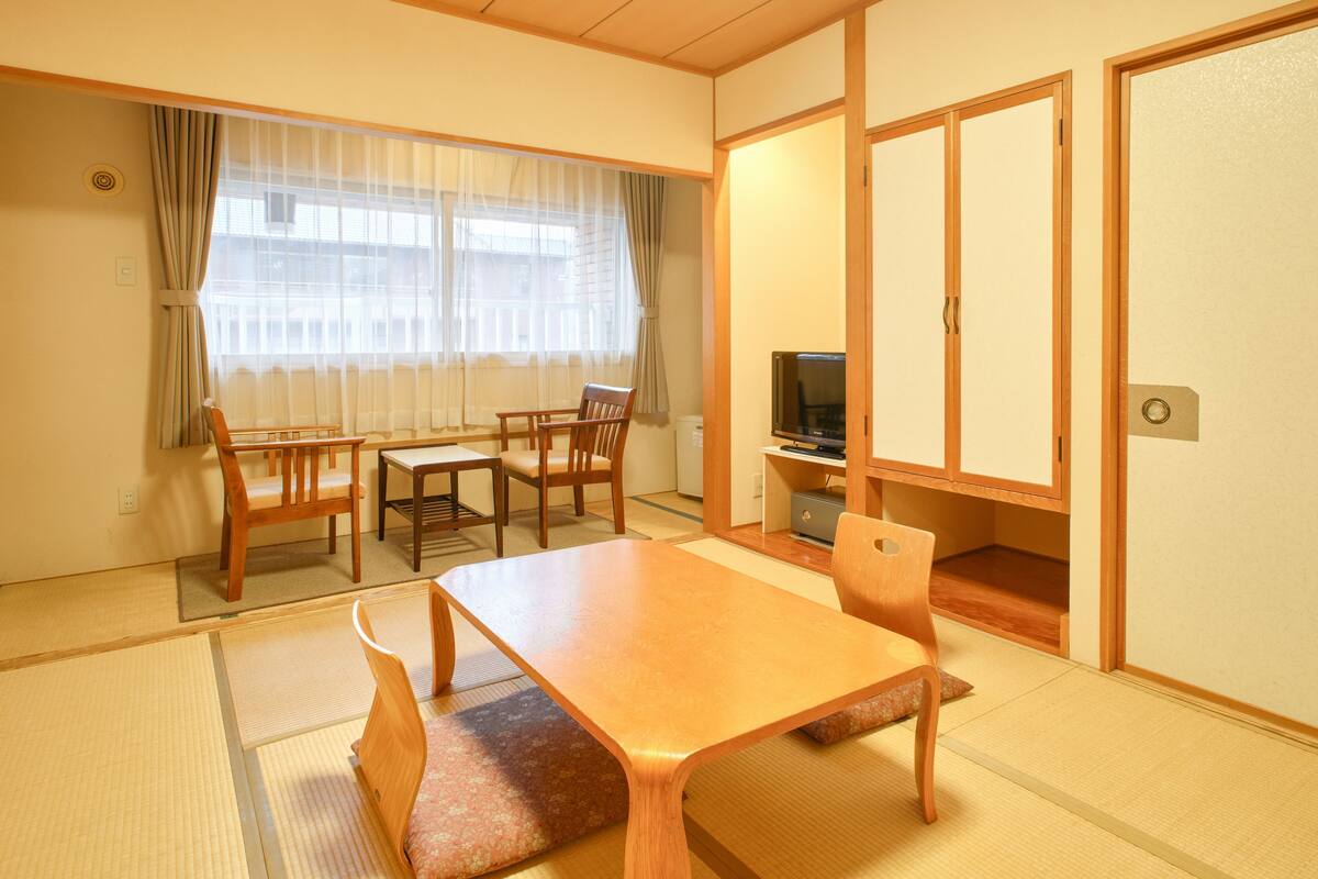 Ein traditioneller japanischer Raum mit Tatami-Böden, einem niedrigen Tisch, Holzstühlen, einem Fernseher und Schiebetüren.