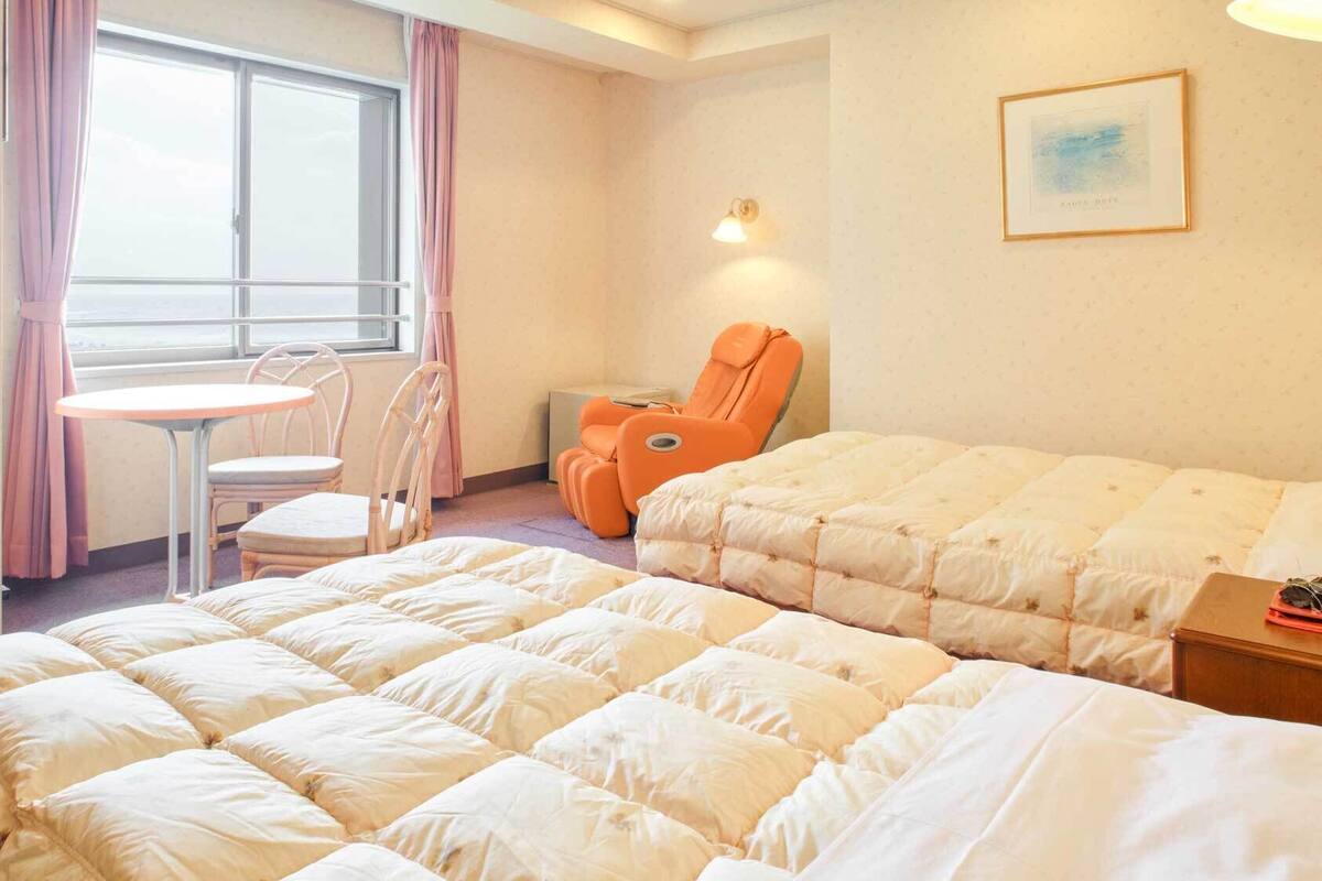 Ein Hotelzimmer mit zwei Betten, einem kleinen Tisch mit Stühlen, einem Massagesessel, einem Fenster mit Vorhängen und einer Wandlampe.