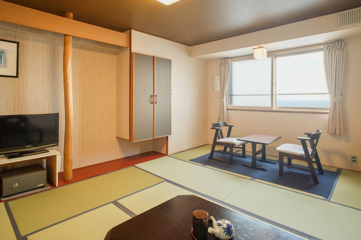 Ein Zimmer mit Tatami-Böden, einem Fernseher, einem Schrank, einem Tisch mit Stühlen und einem Fenster mit Vorhängen.