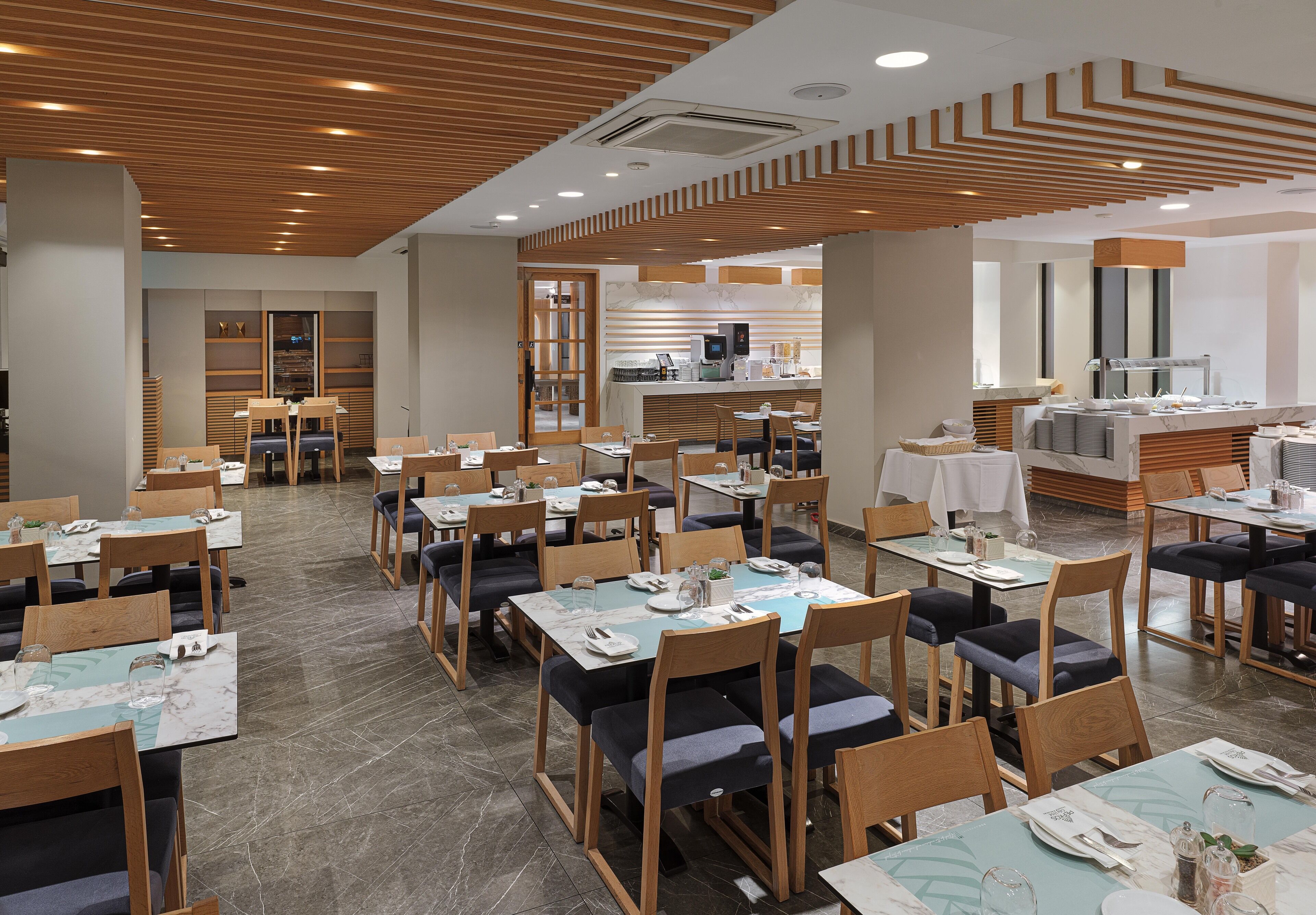 Un restaurante moderno con paneles de techo de madera, mesas de mármol y sillas acolchadas.