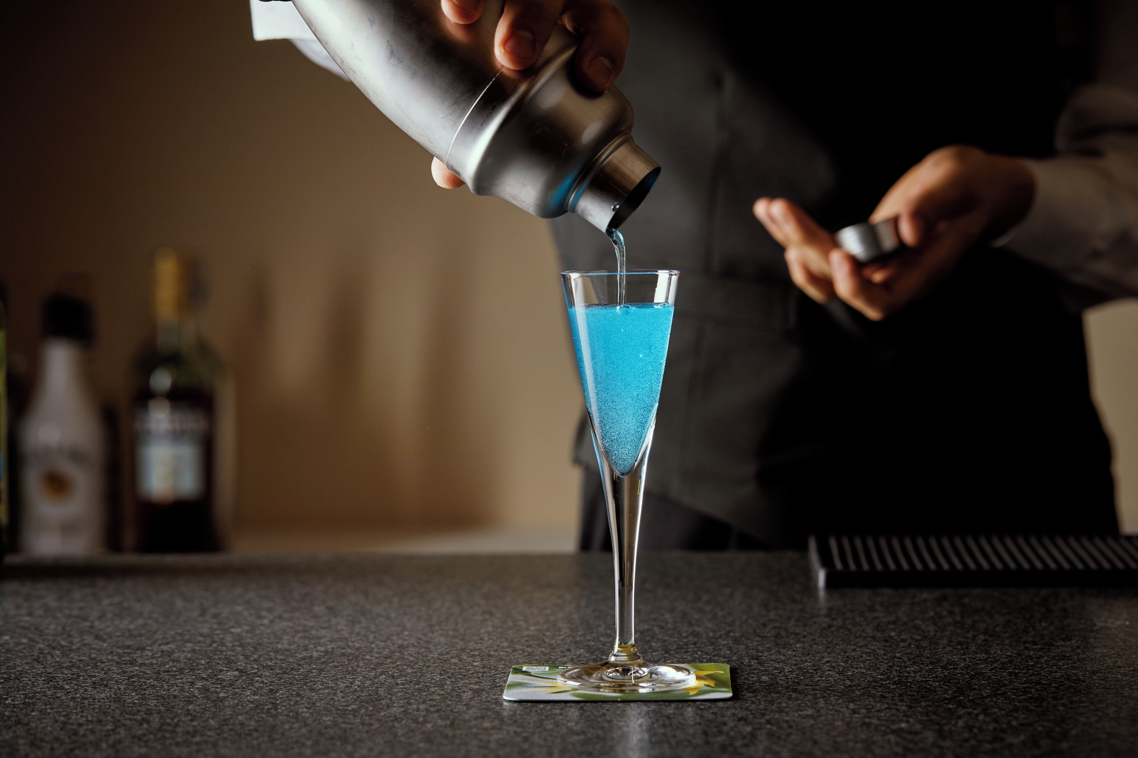 Um bartender servindo um líquido azul em uma taça de martini sobre o balcão do bar.