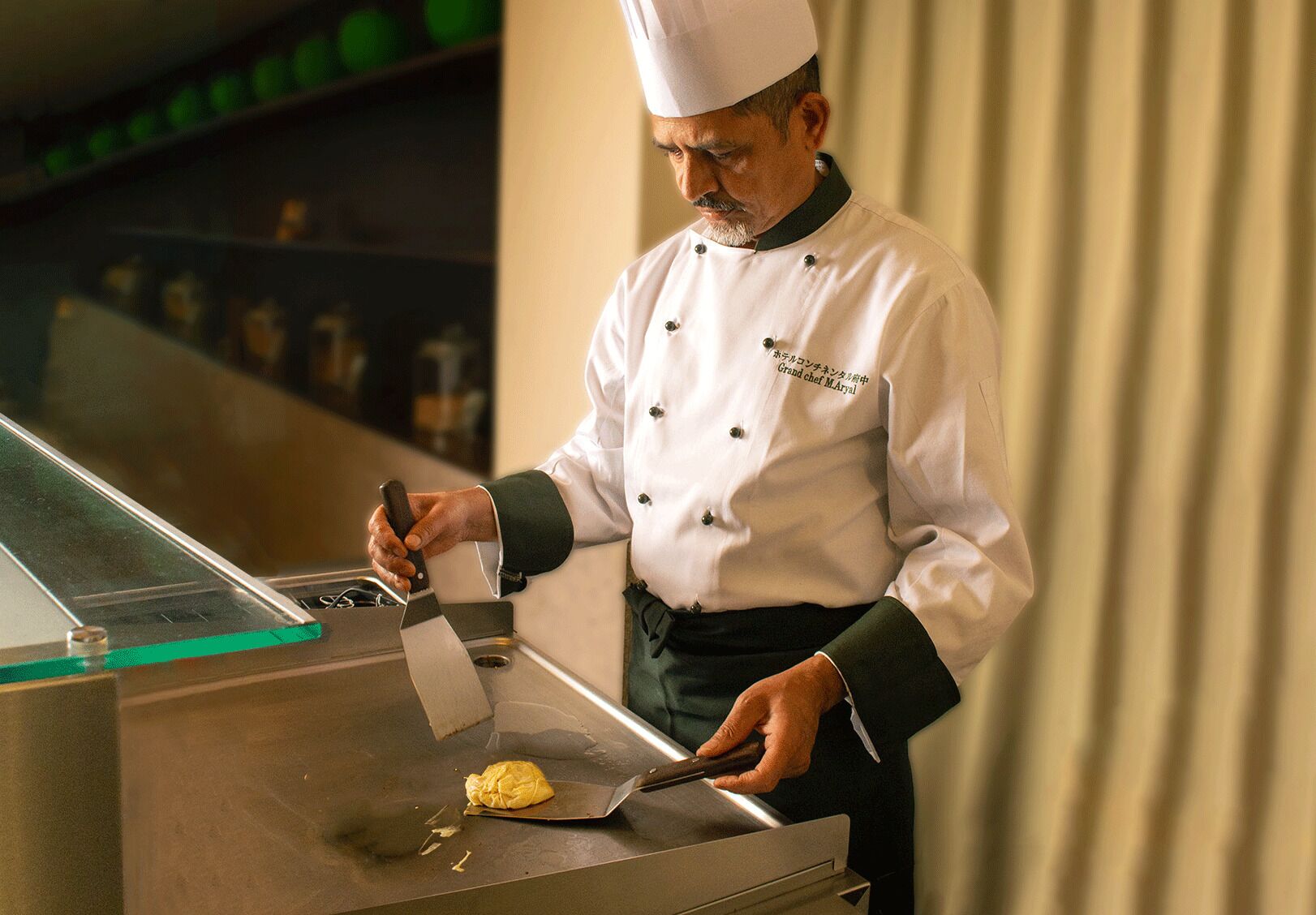 A chef in a white uniform with a black apron, using a spatula to cook on a stainless steel surface.