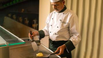 A chef in a white uniform with a black apron, using a spatula to cook on a stainless steel surface.