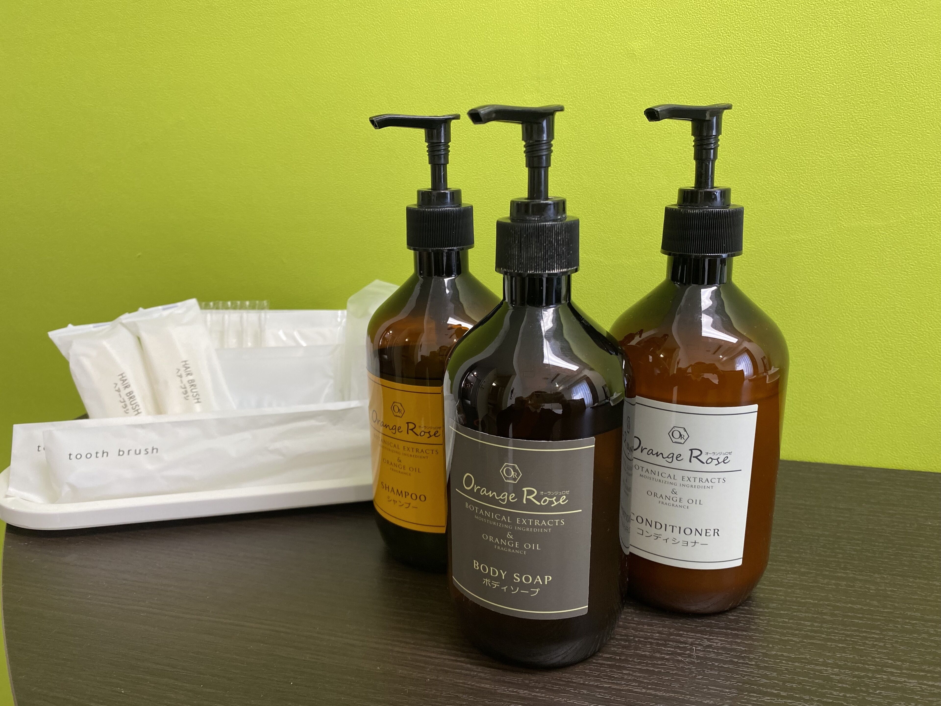 Tres botellas de jabón y champú corporales Orange Rose, una bandeja blanca con paquetes de cepillo de dientes y una pared verde al fondo.
