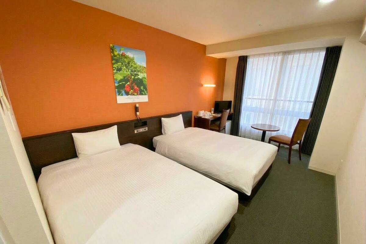 Habitación de hotel con dos camas, un escritorio, una silla, una mesita, un cuadro en la pared y una ventana con cortinas.