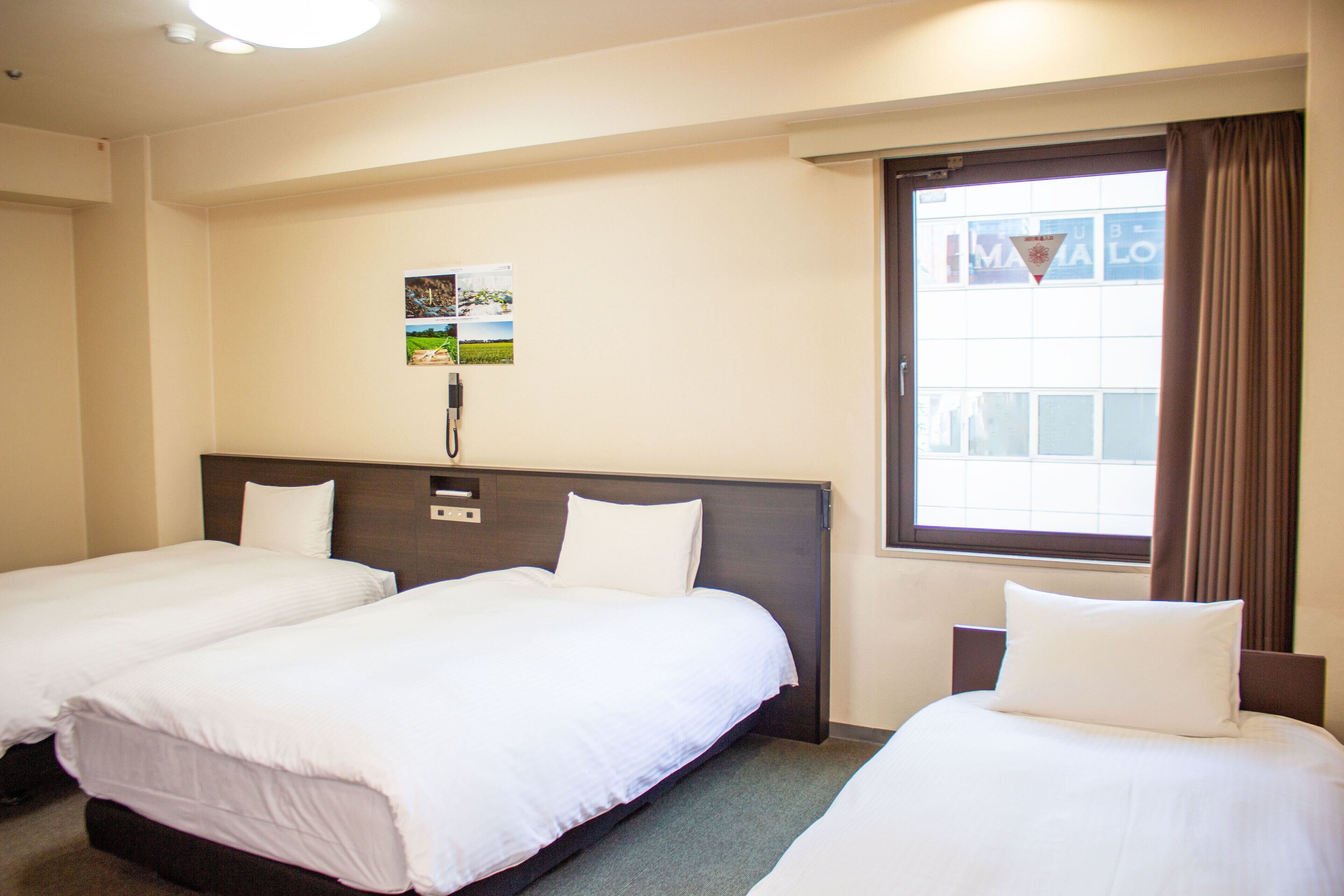 A hotel room with two single beds, a large window with curtains, and a wall-mounted phone.