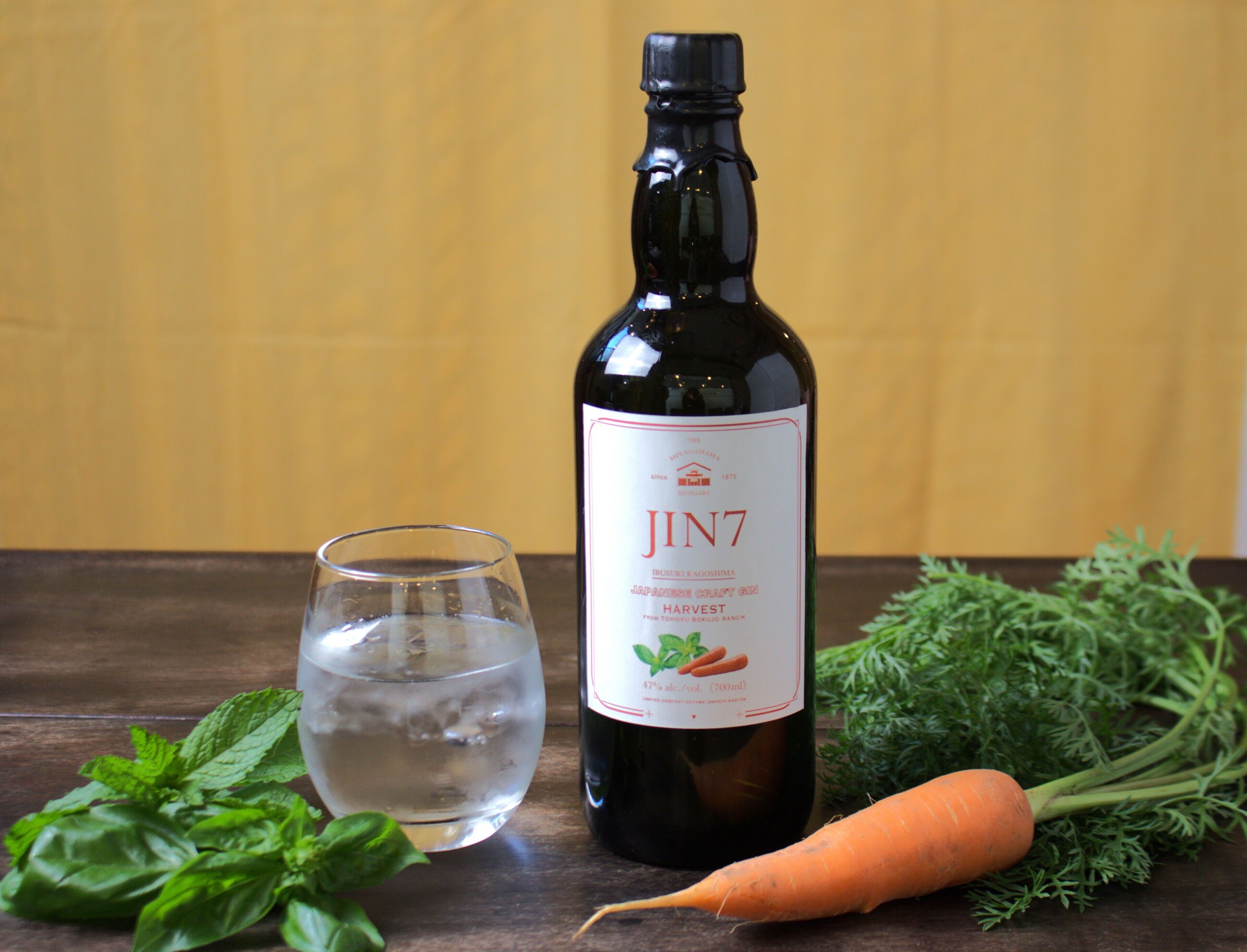 A bottle of Jin7 Japanese grape wine, a glass of water, fresh basil, and a carrot on a wooden surface.