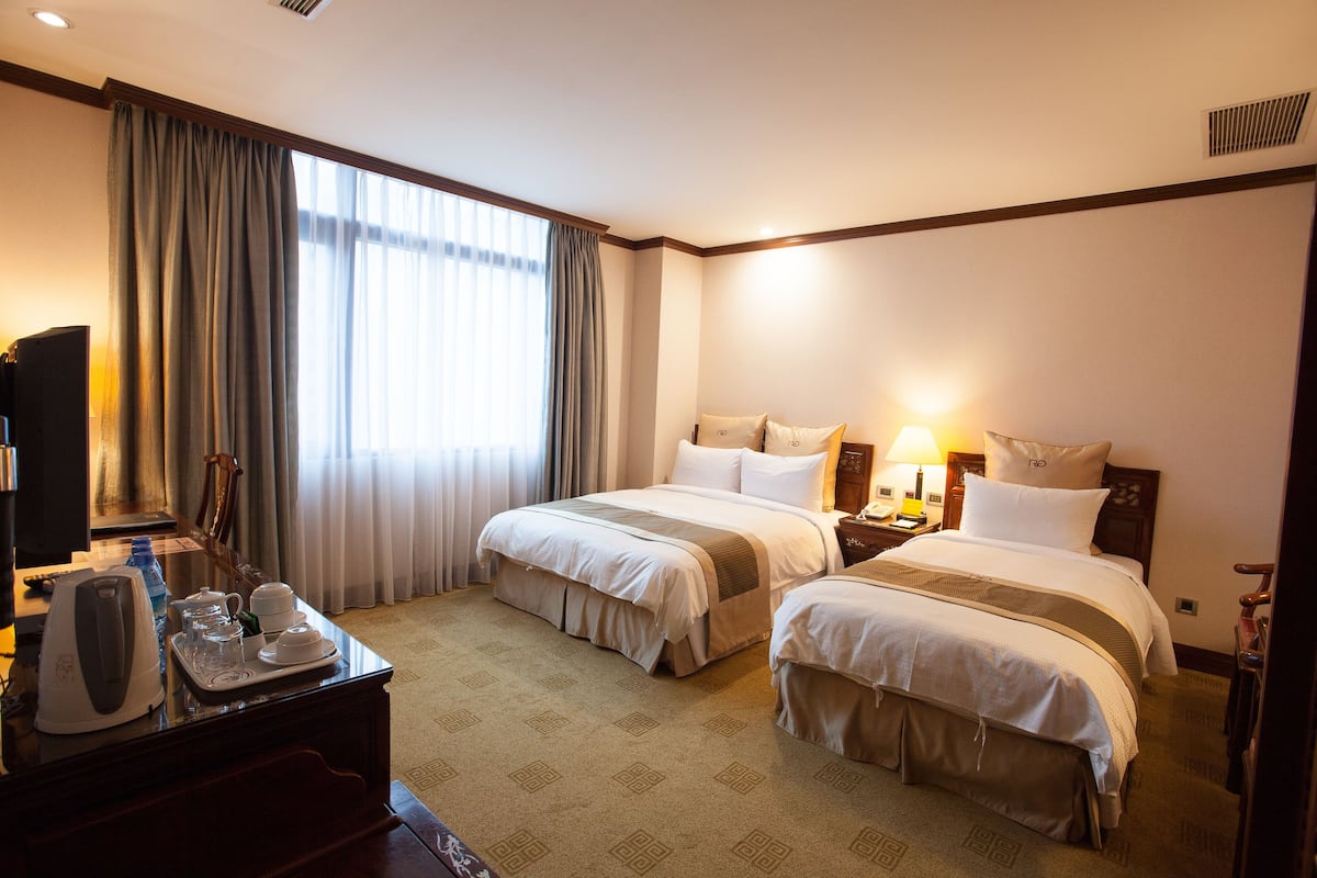A hotel room with two beds, a desk with a kettle and cups, a TV, and a window with curtains.