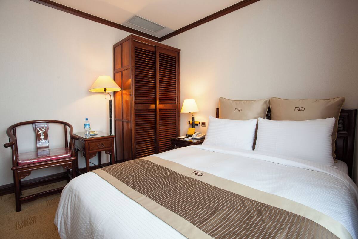 A hotel room with a large bed, a wooden wardrobe, a desk with a lamp, and a chair.