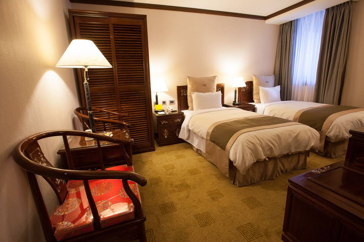 A hotel room with two beds, a desk, a chair, and a lamp.