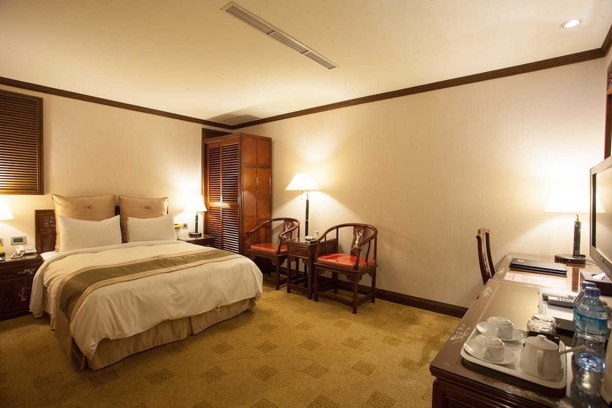 A hotel room with a bed, two chairs, a desk with a lamp, and a water bottle.