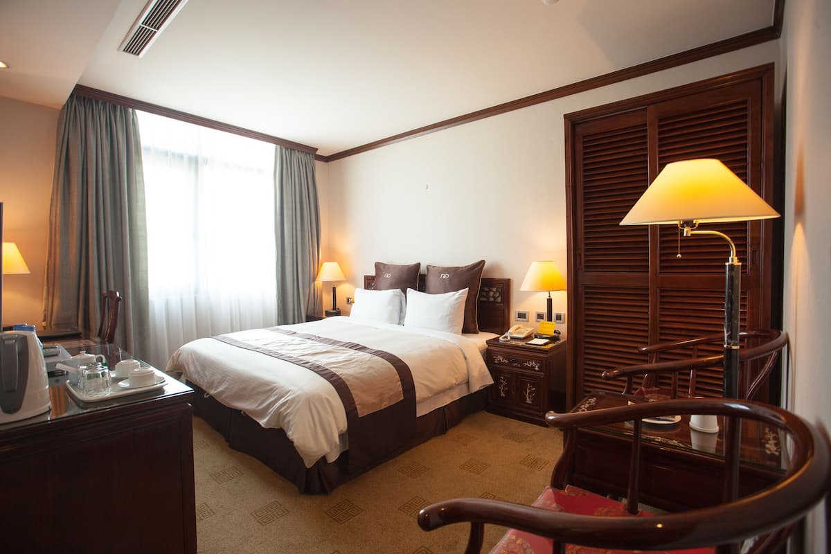 A hotel room with a large bed, bedside lamps, a desk with a telephone, and a view of the outside through a window with curtains.