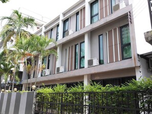 A multi-story building with air conditioning units on the exterior, a balcony with a railing, and a fence with greenery. - Varinda Hostel (Bangkok)