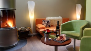 Un salon chaleureux avec un poêle à bois, un fauteuil vert, un canapé recouvert d’un plaid moelleux et une table sur laquelle se trouvent une bouteille de vin et un panier de fleurs.