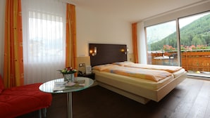 Une chambre d’hôtel avec un lit, une table en verre ornée d’un vase de fleurs, un fauteuil rouge et une vue sur un jardin, visible à travers une grande fenêtre.