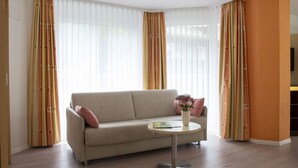 Un salon avec un canapé beige, une petite table ronde ornée d’un vase de fleurs et des rideaux à motifs rayés.