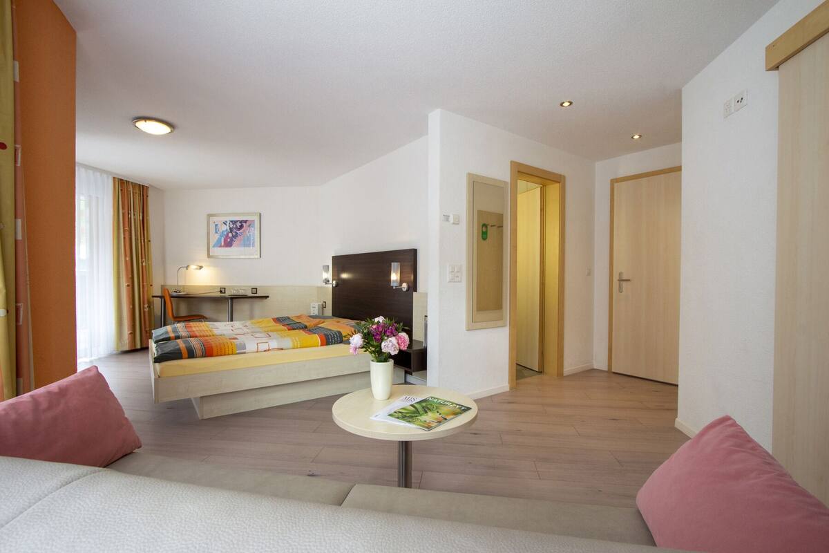 A hotel room with a bed, a small desk, a chair, a round table with a magazine, and a flower vase.