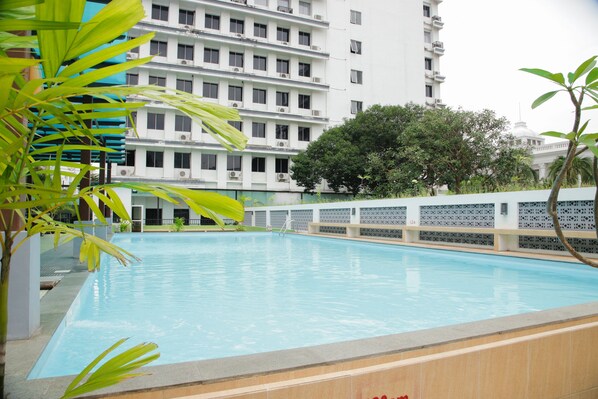 A swimming pool with a tiled edge, surrounded by a low wall and palm trees. In the background, there is a multi-story building with balconies and air conditioning units. - Grand Inna Medan (Medan)