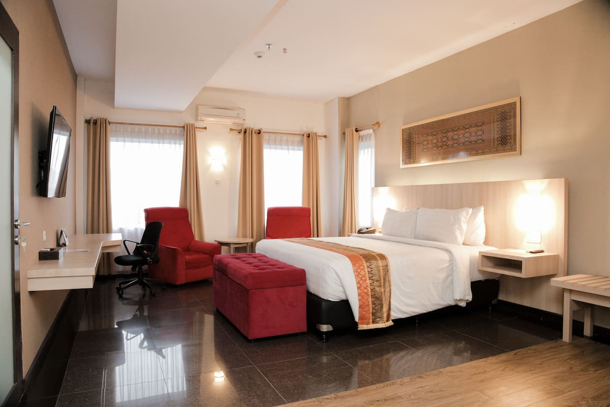 A hotel room with a bed, a desk, a red chair, a red ottoman, and a television on the wall.