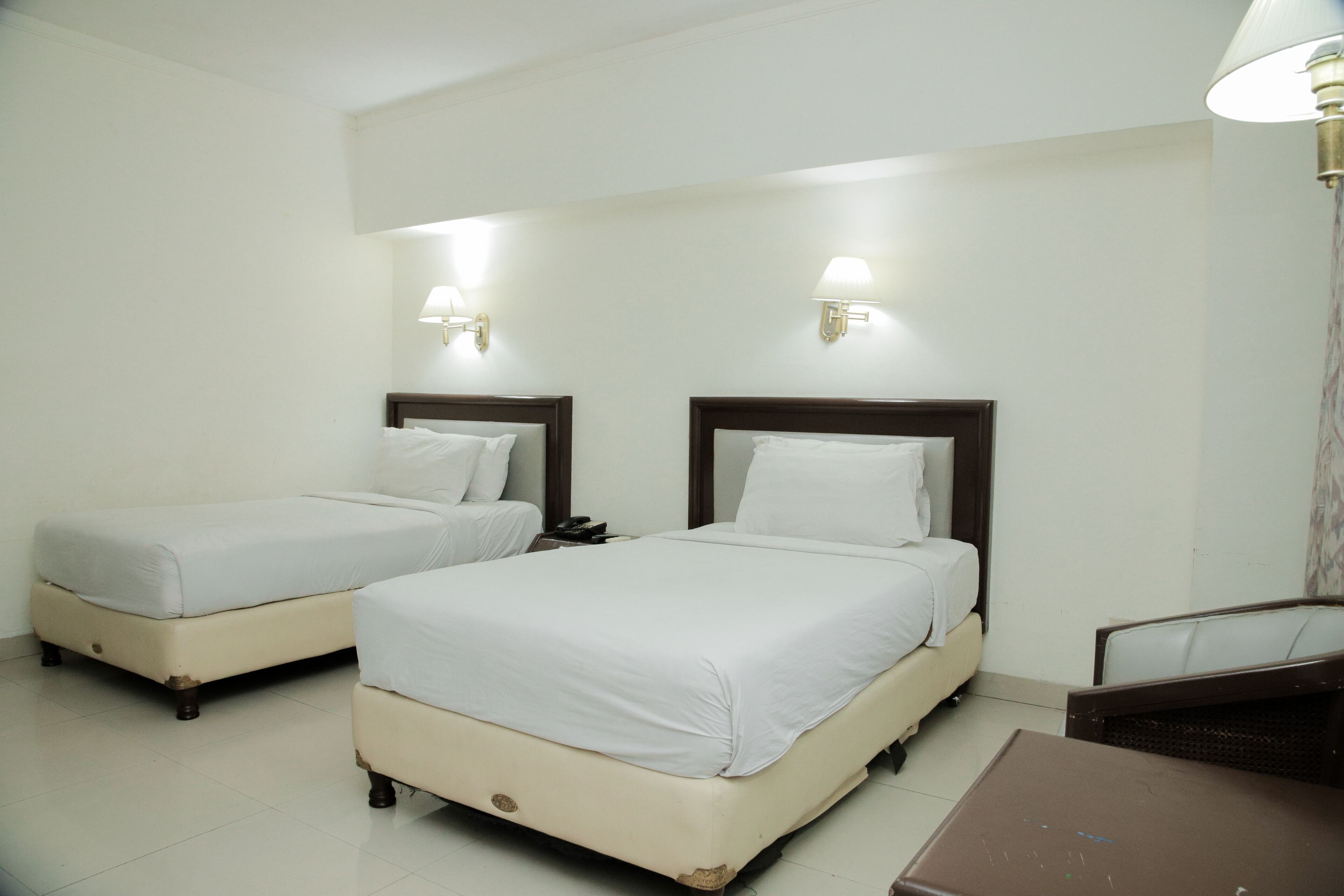 A hotel room with two beds, each with a wooden headboard and white bedding. There are wall-mounted lamps and a table with a chair.