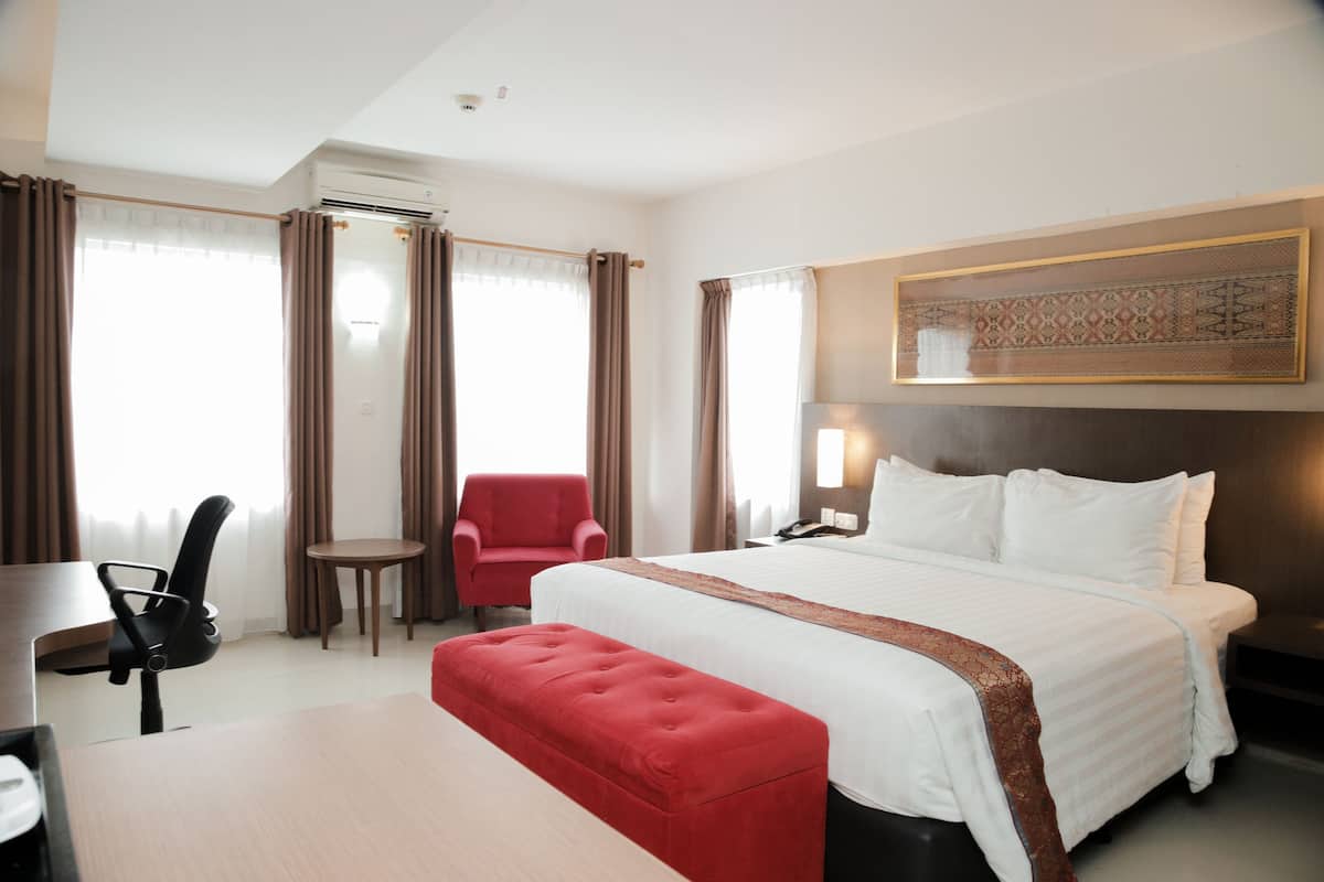 A hotel room with a bed, a desk, a chair, and a red ottoman.