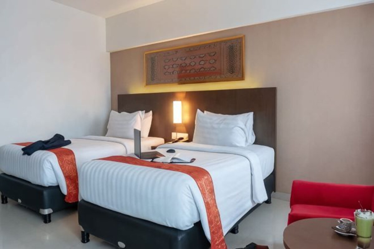Habitación de hotel con dos camas, un sofá rojo, una mesita con una bebida y una lámpara fijada a la pared.