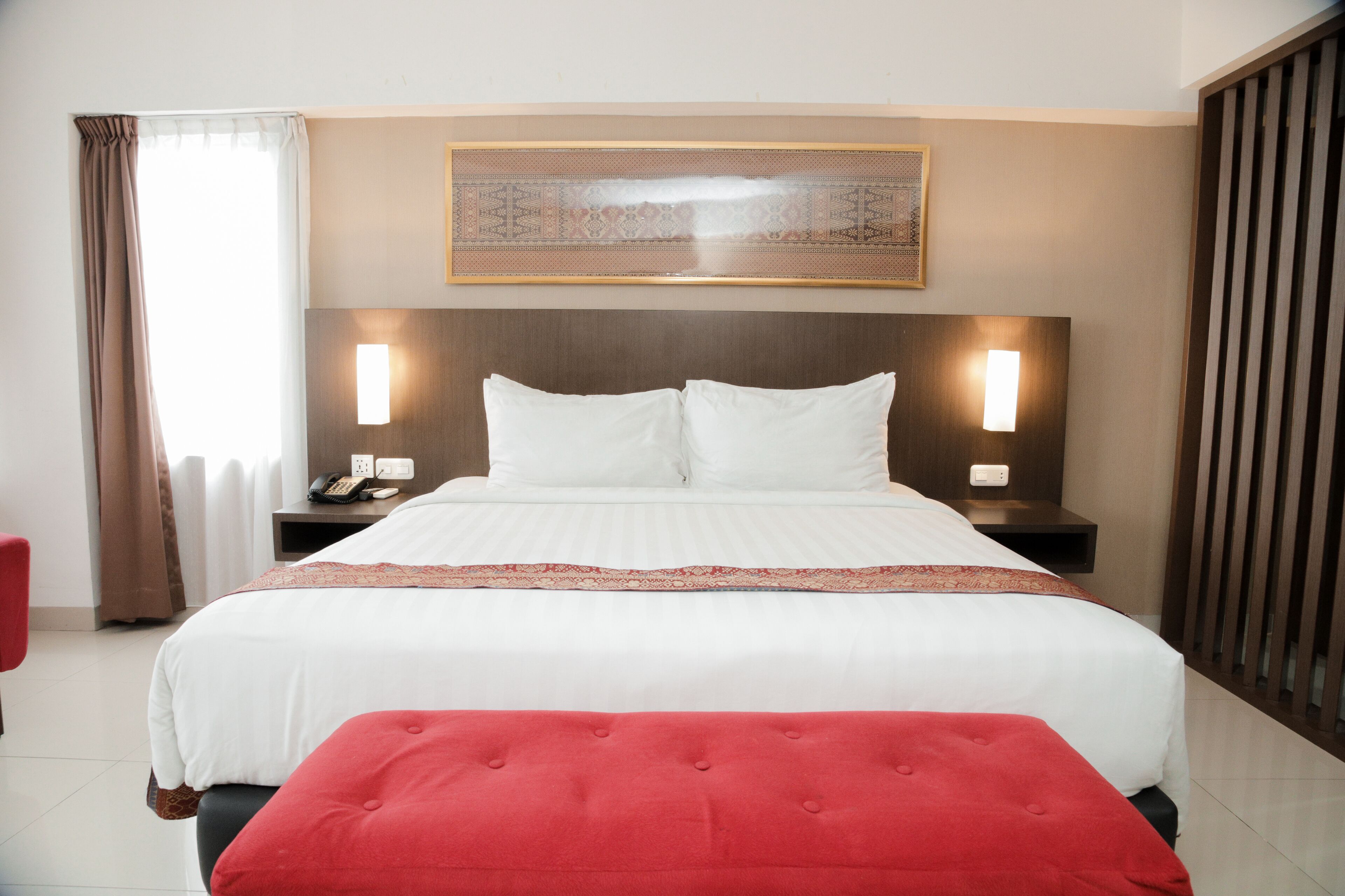 A hotel room with a large bed, two bedside tables with lamps, a red ottoman, a window with curtains, and a decorative wall hanging.
