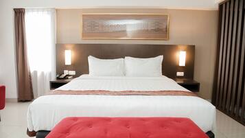 A hotel room with a large bed, two bedside tables with lamps, a red ottoman, a window with curtains, and a decorative wall hanging.