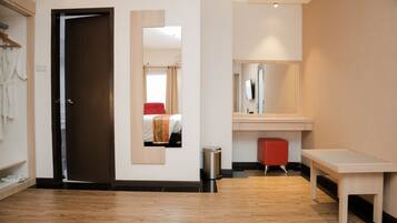 A hotel room with a wooden floor, a bed with a red accent, a small red stool, a wooden bench, a mirror, and a trash bin.