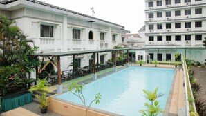 Une piscine entourée d’un espace extérieur couvert, aménagé avec des tables et des chaises, jouxtant un bâtiment blanc doté de nombreuses fenêtres et de balcons.