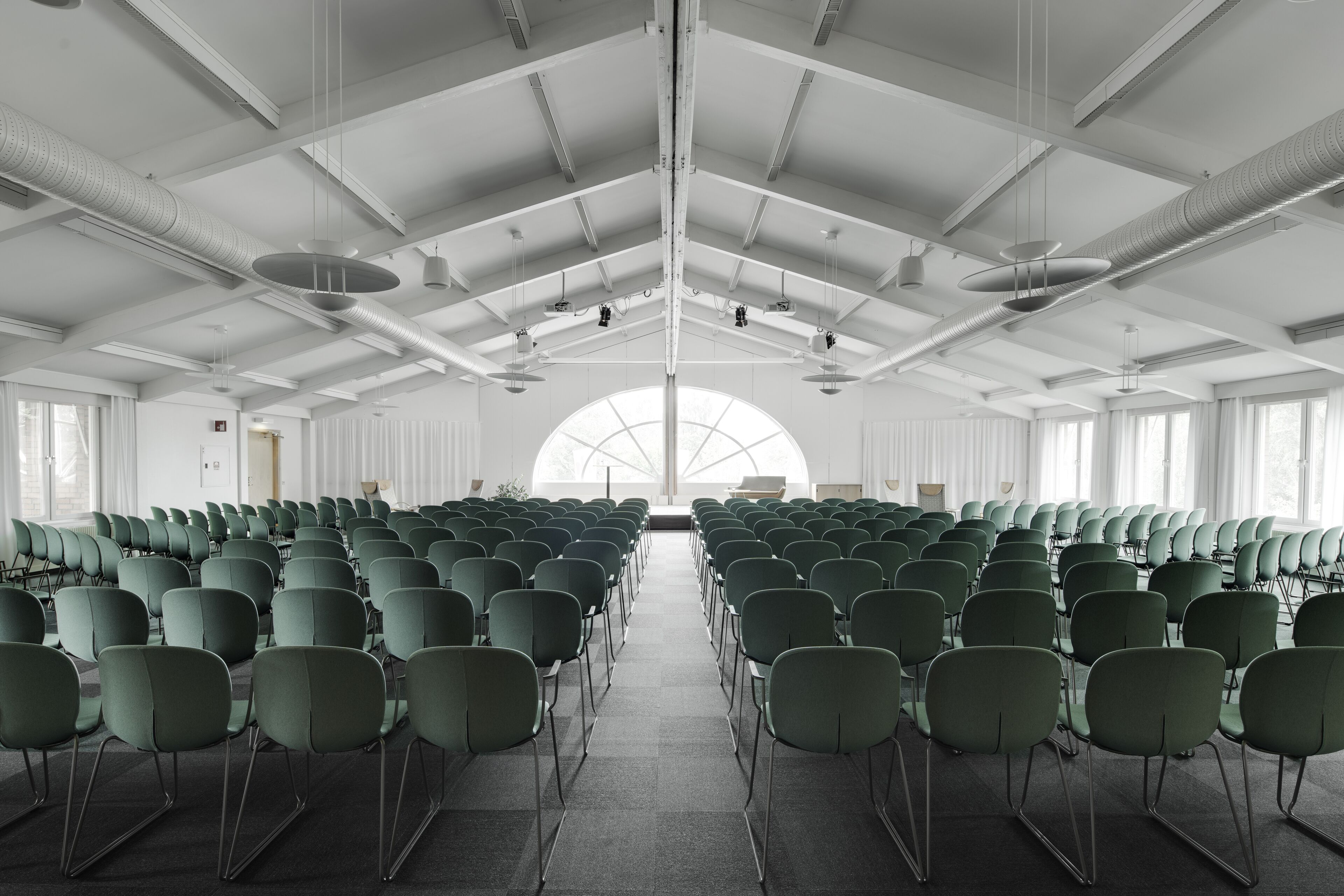 Un salón de conferencias con filas de sillas, un techo alto con vigas a la vista y ventanas amplias que dejan entrar luz natural.