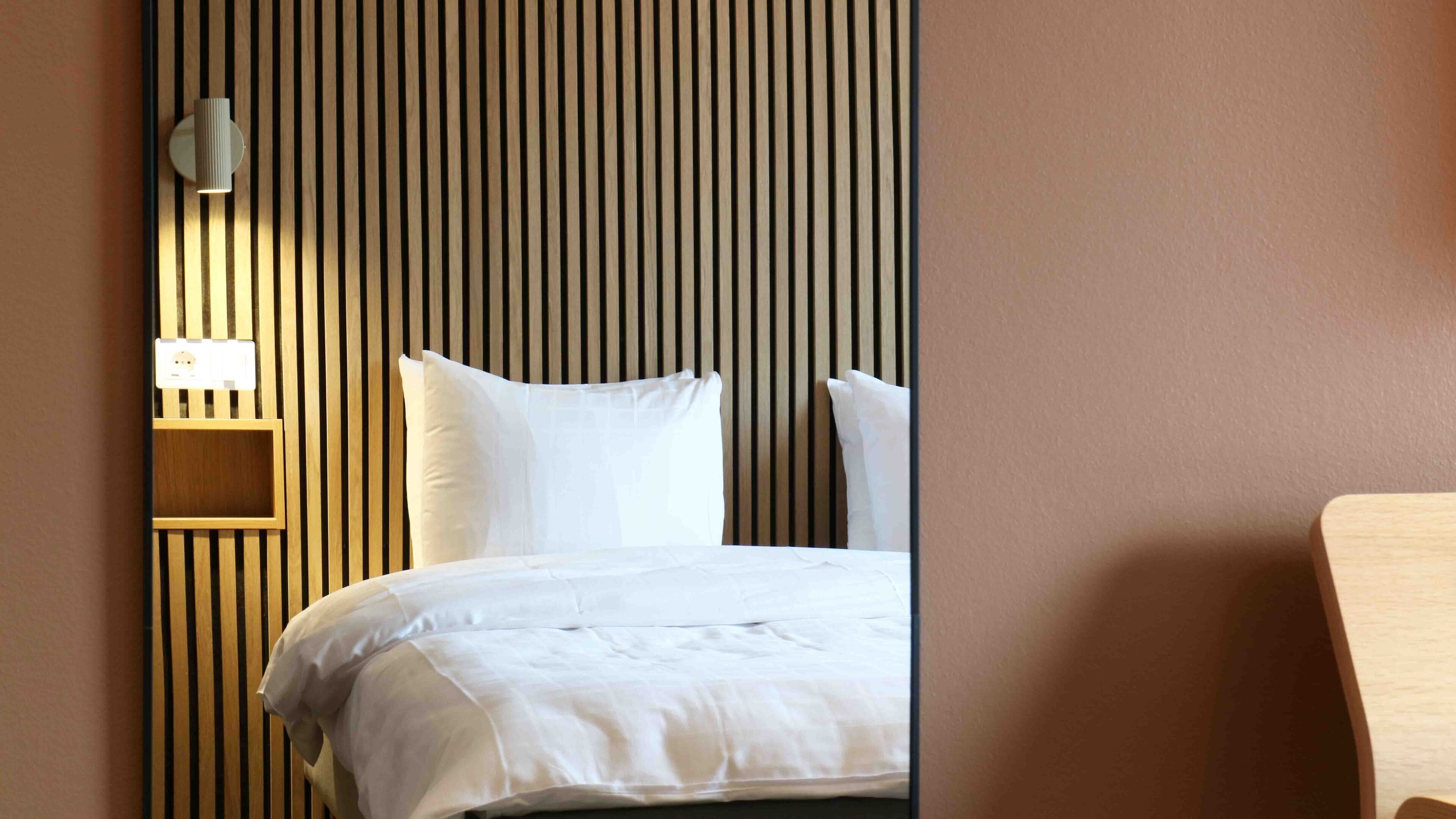 A hotel room with a bed, white bedding, a wooden slat headboard, a bedside table, and a wall-mounted lamp.