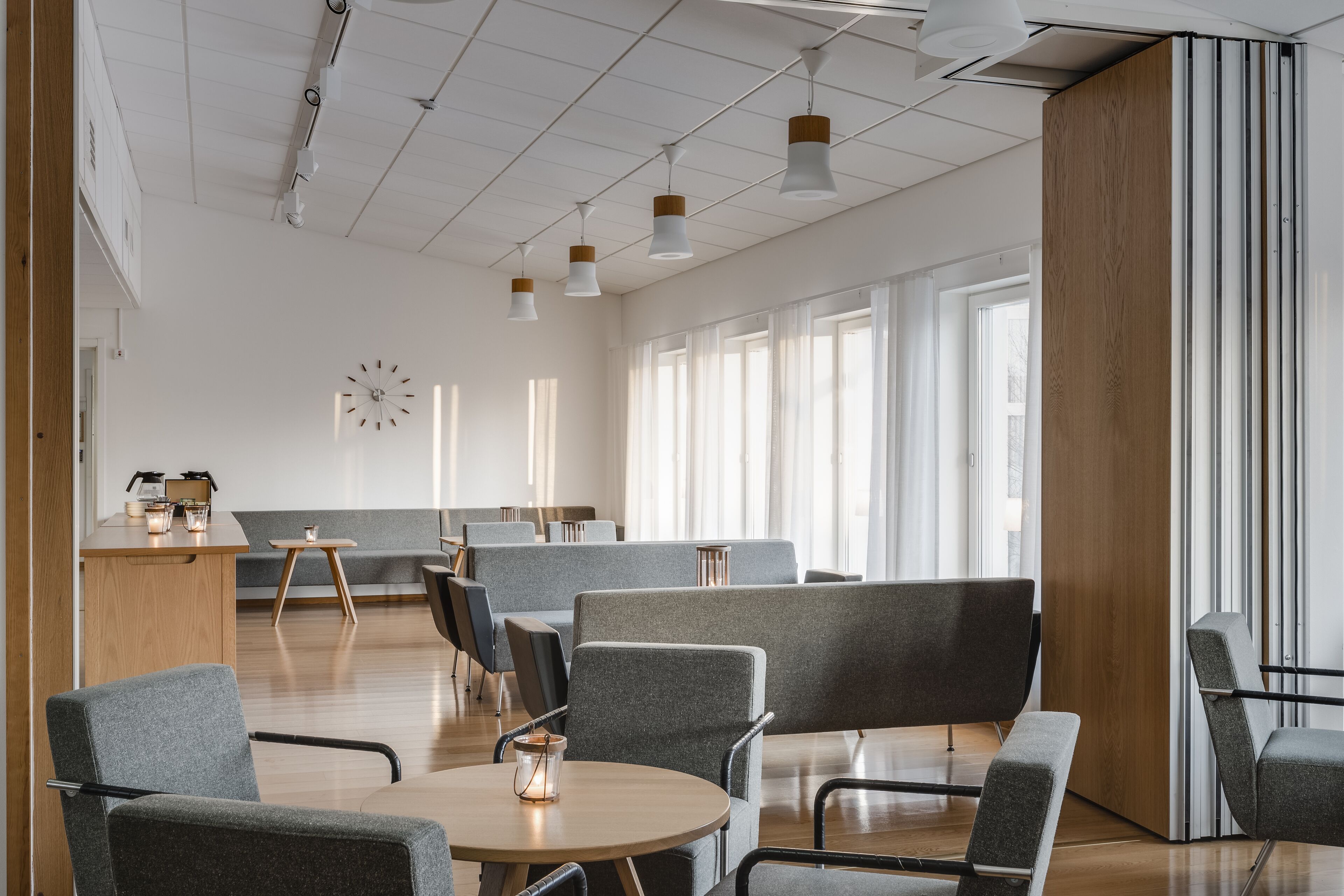 A modern, well-lit room with grey upholstered chairs, a wooden table, and a large window providing natural light.