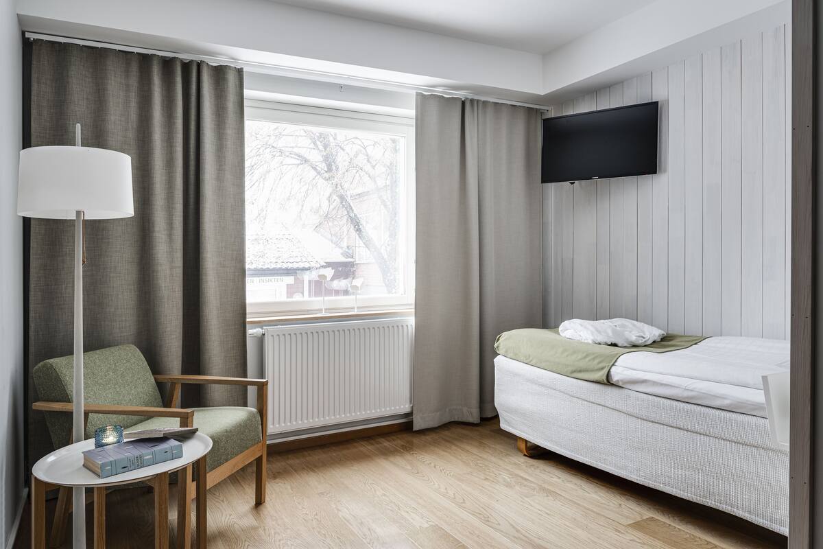 Ein Hotelzimmer mit einem Bett, einem Sessel, einem kleinen Tisch, einer Lampe, einem Fernseher und einem Fenster mit Vorhängen.