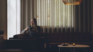 Une personne est assise sur un canapé dans une pièce faiblement éclairée, avec une grande fenêtre et un luminaire suspendu.