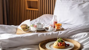 Un lit bien fait, avec du linge de lit blanc, une tête de lit en bois, une table de chevet sur laquelle se trouve un plateau de petit-déjeuner, une bouteille de jus d’orange, une tasse et une petite assiette avec de la nourriture.