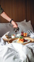 A breakfast setting with a person pouring a drink into a cup, a sandwich, and a plate with food items on a wooden tray.